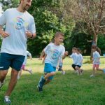A group of kids running with an instructor during outdoor sports activities on a sunny day in a park.